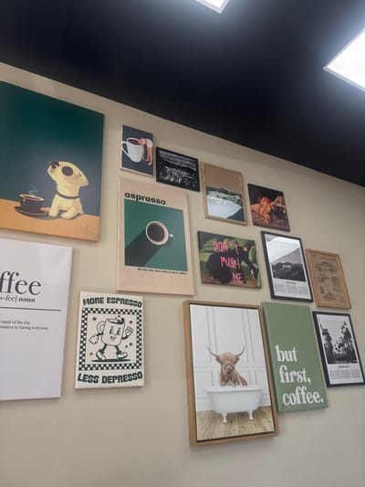 The image showcases a light-colored wall functioning as a gallery, displaying various framed and unframed artworks, predominantly coffee-themed. This commercial establishment is located in Glendale, United States. A black ceiling with two recessed square light fixtures is visible at the top. The artworks are arranged in an informal grid.
**Key pieces include:**
**Top Left:** A large, unframed canvas featuring an illustrated yellow dog in sunglasses beside a steaming coffee cup, against a dark teal and orange background.
**Top Row (smaller items):** A vertical framed print depicts a person draped over a white coffee cup. Adjacent to it, a horizontal framed black and white cityscape or landscape photo. Next, a horizontal framed print shows a vintage sports car's side profile. The row concludes with a horizontal framed still life of croissants, coffee beans, and a coffee mug in warm tones.
**Middle Row:** A large vertical unframed cream-colored poster declares "espresso" and "the day only starts after a cup of coffee," illustrating a white coffee cup on a dark teal background. Beside it, a horizontal unframed canvas features a figure on a green surface with pink text "DON'T PUSH ME." Further along, a vertical framed print captures the "HOLLYWOOD" sign and hills in black and white. A sepia-toned technical drawing of a vintage car in a vertical framed print completes the row.
**Bottom Row:** The far left holds a vertical unframed canvas with a dictionary-style definition of "coffee," including "The best meal of the day. A delightful alternative to hating everyone." Next, a vertical unframed canvas presents a cartoon coffee cup with "LESS DEPRESSO MORE ESPRESSO" on a checkered border. A vertical framed canvas displays a Highland cow in a bubble bath. To its right, a solid sage green unframed canvas reads "but first, coffee." The rightmost piece is a vertical framed black and white photograph of a tree-lined street with text, possibly "BEVERLY HILLS."