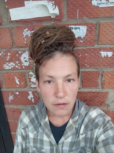 A close-up selfie shows a person with dreadlocks piled on top of their head, standing against a brick wall with torn posters. The person is wearing a gray and white plaid shirt over a dark shirt. The background consists of red bricks with various pieces of torn paper adhered to them. The lighting is natural and appears to be from the front, casting soft shadows. The overall scene suggests an urban outdoor setting.