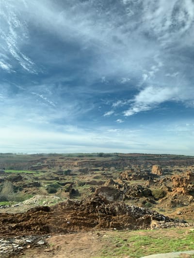 The image captures an expansive, eroded landscape in Sidi Slimane Moul Al Kifane, Morocco, under a bright blue sky partially covered by cirrus and scattered cumulus clouds. The scene is dominated by undulating terrain featuring numerous gullies, steep banks, and exposed layers of reddish-brown and orange-brown earth, indicative of significant soil erosion. Patches of vibrant green vegetation, including grasses and shrubs, are visible within depressions and across less eroded areas. In the distant background, a strip of cultivated land with organized rows of trees or crops is discernible along the horizon. The immediate foreground consists of a mound of dark brown, disturbed soil, with small fragments of light-colored debris scattered upon it, and a section of green grass to the right. No individuals, animals, or direct human actions are observed within the frame.