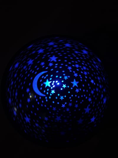 A close-up shot shows a dark sphere with blue star and crescent moon patterns on its surface. The sphere appears to be emitting light, giving the patterns a glowing effect. The background is entirely black, emphasizing the illuminated sphere. There is no indication of a location in Borino, Bulgaria, or any other information about people, subjects, objects, actions, or interactions beyond the described sphere and background.