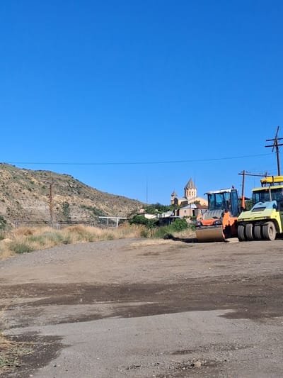 In the city of Vayk, Armenia, under a clear blue sky, a construction site is depicted. The foreground is a rough, dusty dirt and gravel lot. In the middle ground, two road rollers are parked. The larger, orange roller is positioned to the left, and a yellow roller is to its right. Behind the rollers, a cluster of buildings is visible, including a church with distinct spires. The background features a barren, rocky hillside sparsely covered with dry vegetation. Power lines and poles are also present in the scene. The overall impression is one of an active or recently active construction zone in a semi-arid environment.