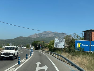 A white 4x4 vehicle is driving on a two-lane road in Olesa de Montserrat, Spain. The road curves to the right and is lined with blue and white bollards. In the background, there is a mountainous landscape with green hills and a clear blue sky. A large signpost indicates directions to "MONISTROL DE MONTSERRAT MANRESA" straight ahead and "La Flora OLESA DE MONTSERRAT ESPARREGUERA" to the right. Further down the road, a large blue billboard displays the text "Sortida L Flor" and an image of an exit ramp. There are other vehicles on the road, including a white van and a bus. The foreground shows the road markings with directional arrows indicating a right turn. The environment appears to be rural and sunny.