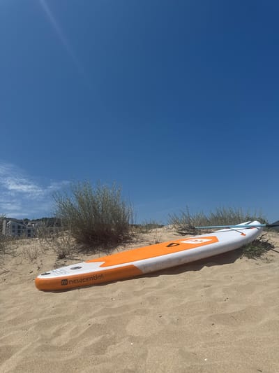 A stand-up paddleboard with an orange and white design lies on a sandy beach. The paddleboard has the brand name "newcential" printed on its side. In the background, sparse dry vegetation and some buildings are visible against a clear blue sky. The scene appears to be in the city of Dyuni, Bulgaria.