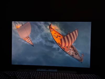 The media file displays a television screen showing an animated scene with two Viking-style ships sailing through a cloudy, dark sky. The ship in the foreground has a large sail with red and white stripes and a wooden deck filled with barrels and what appear to be figures. The ship's prow is shaped like an animal's head. The second ship, further in the background and to the left, has a white sail and also appears to be a Viking longship. Both ships have lit torches or lanterns, suggesting it might be dusk or nighttime. The overall mood of the scene is dramatic and adventurous, with the dark clouds and the ships navigating through them. The image is taken from a low angle, with the bottom of the television screen and a checkered blanket visible in the foreground. There is no visible text in the media file. The location context provided (Sao Thong Hin Town Municipality, Thailand) does not appear to be directly depicted or suggested by the content of the media file itself.