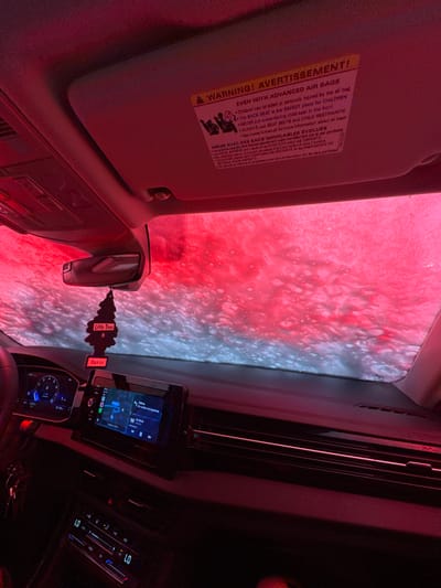 The image depicts the interior of a vehicle, observed from a low angle, while actively undergoing an automated car wash in Roselle, United States of America. The exterior view through the windshield and sunroof is dominated by thick, red-tinted foamy water and numerous white bubbles, indicative of a washing cycle. The red light emanating from the car wash facility heavily illuminates the car's interior.
Inside the vehicle, the dashboard is visible, featuring a digital instrument cluster on the left displaying numerical values and a blue graphic accent. A central infotainment screen shows a navigation interface with a map, a turn instruction in Spanish ("Da vuelta a la izquierda" - Turn left), and music playback information ("Un Beso"). Below the screen, the climate control panel displays "LO" on both driver and passenger sides. A black "Little Trees" air freshener, labeled "Black Ice," hangs from the rearview mirror, which is centrally positioned. The passenger-side sun visor is flipped up, revealing a yellow and white warning sticker with text in English and French regarding "ADVANCED AIR BAGS" and "CHILD RESTRAINTS," emphasizing the back seat as the safest for children. A portion of the steering wheel is visible on the left, and a set of keys hangs near the lower left dashboard area. No individuals are visible within the frame.