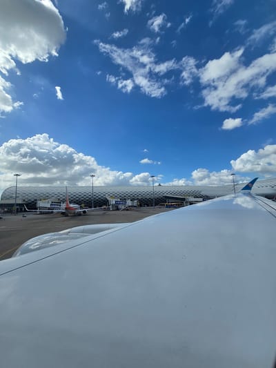 The image captures an aerial view from an airplane wing, showcasing a modern airport terminal under a bright blue sky with scattered white clouds. The terminal features a distinctive, patterned facade with honeycomb-like structures. Several airplanes are visible on the tarmac, connected to jet bridges. The wing of the aircraft in the foreground is prominent, reflecting the sky and clouds. The location is identified as Bao'an District, China.