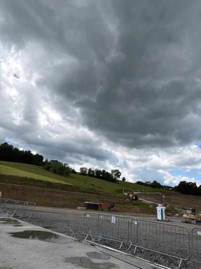 The image depicts a construction site in a rural area with a hill in the background. The sky is filled with dramatic, dark storm clouds. In the foreground, there are several metal safety barriers and puddles of water on a concrete surface, indicating recent rain. The construction site itself is characterized by piles of gravel, a portable toilet, and heavy machinery, including an excavator and a bulldozer. A large black container is also visible. The background features a grassy hillside dotted with trees and a few buildings, suggesting a residential area or scattered houses. The overall impression is one of a vast open space undergoing significant development under an impending storm.