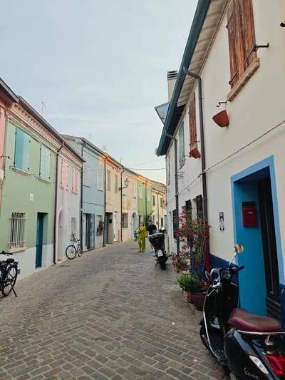 The image depicts a charming, narrow cobblestone street lined with brightly colored houses in Rimini, Italy. The buildings feature pastel facades in shades of pink, green, blue, yellow, and cream, with contrasting shutters and doors. On the right side of the street, a white building with a vibrant blue doorway and a red mailbox is prominent. A black Vespa scooter with a burgundy seat is parked in front of this building.
In the middle ground of the street, a woman wearing a bright yellow jumpsuit walks away from the camera. Another person, partially obscured, is walking behind her. A bicycle is parked on the left side of the street. The street is paved with grey cobblestones, and potted plants with red flowers add a touch of color.
The scene appears to be set in the late afternoon or early evening, indicated by the soft, warm light casting subtle shadows and the overall gentle illumination. The sky is a pale blue with wispy clouds, suggesting fair weather. The architecture, including the shutters, window designs, and the style of the houses, is characteristic of Italian coastal towns. The presence of bicycles and scooters suggests a pedestrian-friendly and leisurely environment. The overall mood is peaceful and serene, evoking a sense of a quiet, picturesque Italian town.
A license plate is visible on the back of the Vespa, reading "P 57122". A small black and white sign with the text "D.I.Y." is affixed to the white wall on the right.