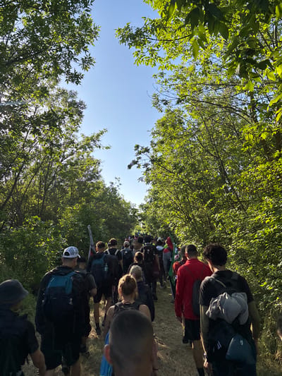 A group of people is walking on a path surrounded by trees. The scene is shot from a low angle, looking up at the trees and sky. Most of the people are wearing backpacks. The location is Lipnitsa, Bulgaria.