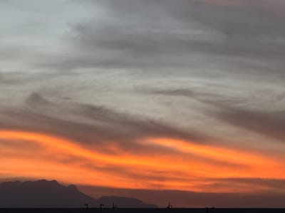 This is a landscape image captured at either sunrise or sunset, showcasing a vibrant sky over a silhouetted landform in San Francisco Ocotlán (Ocotlán), Mexico. The sky dominates the frame, displaying a dramatic color gradient. The upper portion of the sky is a muted gray and off-white, streaked with delicate, wispy clouds that transition into horizontal bands of brilliant orange and red closer to the horizon. These warm hues suggest the golden hour light illuminating the cloud formations.

Below the colorful sky, a dark, undulating silhouette of mountains or hills stretches across the middle ground, their jagged peaks outlined against the bright light. In the immediate foreground, a dark, flat horizontal line suggests a structure like a rooftop or a low wall. From this dark foreground, several small, dark, pipe-like or vent-like objects protrude upwards at irregular intervals. There are no people or explicit activities visible in the frame. The overall impression is one of natural beauty and tranquility, highlighted by the striking colors of the sky. No text is visible in the image.