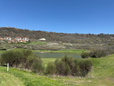 This media file captures a wide, elevated view of a scenic landscape encompassing a golf course, residential buildings, and hills under a clear blue sky.

In the foreground, a vibrant green grassy slope is prominent, interspersed with dense, brushy bushes that appear to be leafless or have sparse foliage, possibly indicative of early spring or a specific shrub type. A white marker post stands on the left.

The midground features a tranquil body of water, likely a lake or pond, with an olive-green hue, forming part of a golf course's water hazard. Beyond the water, manicured green fairways and what appear to be white sand bunkers are visible.

The background consists of rolling hills covered in a mix of bare deciduous trees and shrubs, suggesting either late winter or early spring. Several residential or resort buildings with white facades and distinctive reddish-orange tiled roofs are nestled on these hillsides. A cluster of these buildings is situated on the left slope, while a more isolated, larger white building stands atop a distant hill to the right. Winding paths or roads can be seen traversing the hills.

The image depicts a bright, sunny daytime scene with a vast, clear blue sky. No specific activities or events are actively taking place, but the presence of the golf course implies recreational use. There is no discernible text visible in the image. The setting suggests a well-maintained resort or residential area in a natural, hilly environment, consistent with the provided location context of Bozhurets, Bulgaria.