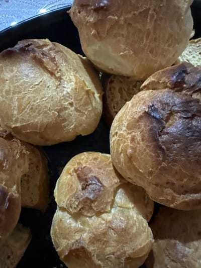 A close-up shot displays a collection of freshly baked pastries, likely cream puffs or profiteroles, arranged on a dark platter. The pastries are golden brown, with some showing darker, caramelized spots, indicating they are well-baked. Their surfaces have a slightly cracked texture, typical of choux pastry. Some of the pastries are partially obscured by others, creating a layered effect. The lighting is somewhat dim, and the focus is on the texture and color of the baked goods. The image appears to have been taken indoors, likely in a kitchen or dining area, given the close-up view and the setting of food preparation or serving. There are no people visible, and the activity is implied to be the presentation or consumption of these pastries. The overall impression is one of warm, comforting food.