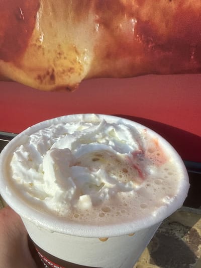 The image shows a close-up, slightly overhead view of a white paper cup filled with a frothy beverage topped with whipped cream. Some pinkish swirls are visible in the whipped cream and liquid. In the background, a blurry, out-of-focus image of food, possibly pizza, is visible against a red surface. The scene appears to be outdoors, with sunlight casting shadows. The cup has a dark brown band near the bottom with some red lettering, but the text is illegible. The photograph was taken in Emerson, United States of America.