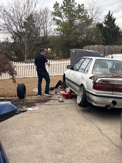 A white Honda wagon is parked on an asphalt driveway with its hood open. A person is working beneath the car using a red jack. A man stands nearby in a black shirt, jeans, and a baseball cap, holding a tool. A tire lies on the ground near a white picket fence. Bare trees and green foliage surround the scene. A blue car's hood is visible in the foreground. The sky is overcast.