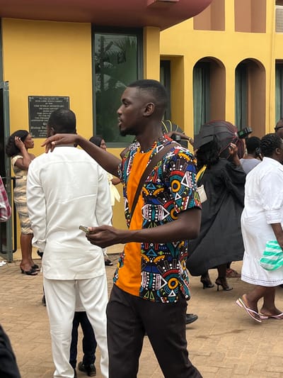 The image captures an outdoor gathering of individuals on a paved area adjacent to a yellow building identified by a wall plaque as the CEM Worldwide Missionary Headquarters in Madina, Ghana. A young adult male, prominently positioned in the foreground slightly right of center, has dark skin, a short fade haircut, and a light beard. He wears a short-sleeved shirt with an orange left side and a multi-colored geometric pattern on the right, dark trousers, and a dark strap crossing his chest. He holds a small, metallic, cylindrical object in his right hand, while his left arm is extended horizontally to the left with an open hand. His head is turned to the right with his mouth slightly open. To his left, a male subject is seen from the back, dressed in a white long-sleeved tunic and matching trousers. Further left, a woman in a light-colored patterned dress has her left hand near her head. In the right background, a person wearing a black academic gown and cap holds a red and black umbrella overhead. To the far right, a woman in a white ruffled dress, carrying a green and white striped bag, is partially visible, wearing pink flip-flops. The building features dark-framed windows with reflections and recessed arched elements on its upper facade. The ground is paved with reddish-brown interlocking pavers.