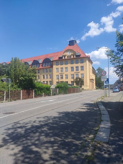 A wide-angle, eye-level shot captures a street scene on a sunny day in Chemnitz, Germany. A large, yellow building with a red tiled roof dominates the left side of the frame, featuring numerous windows and a prominent clock on its facade. Adjacent to the building is a lush green area with trees and a wooden fence. A paved road runs through the foreground, casting dappled shadows from the surrounding trees onto its surface. On the right side of the road, a sidewalk is visible, bordered by stone. Further down the road, other buildings and parked cars can be seen, with trees lining the street.
Traffic signs are visible on the right side of the road, including a speed limit sign indicating "30" and another sign that reads "Schule Mo-Fr 7-16 h," meaning "School Monday-Friday 7 a.m. to 4 p.m." The sky is a bright blue with a few white, fluffy clouds. The overall impression is a peaceful, everyday scene in a European town on a clear, pleasant day. There are no people visible in the frame. The lighting suggests it is daytime, likely mid-morning or afternoon given the shadows.