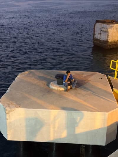 A man sits on a concrete platform by the water in Melaya, Indonesia. He is wearing a blue shirt and jeans, and he is holding a cigarette and a phone. The concrete platform is illuminated by artificial lights, casting long shadows on the ground. The water is dark and calm, with a gentle ripple. In the background, a concrete structure juts out of the water, and a yellow ladder and railing are visible on the right side of the image. The scene appears to be a quiet, contemplative moment by the sea.