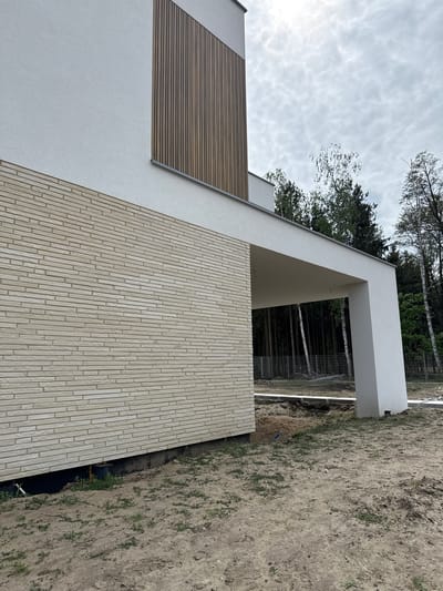 The image shows the exterior of a modern building, likely a house, in Wiązowna, Poland. The building features a minimalist design with a combination of materials: white plastered walls, a section of wood-look slats, and a brick-like facade. A white structural element creates a covered area or entrance. The ground adjacent to the building is bare earth with sparse vegetation. Trees are visible in the background. The sky is overcast.