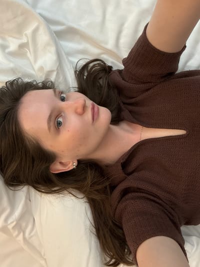 A young woman with long, dark brown hair lies on a white, creased bed. She is wearing a brown, knitted top with a cutout at the chest. Her head is tilted to the left, and she is looking directly at the camera with light blue eyes. Her skin is pale and clear, with a few scattered blemishes. She has a delicate silver chain necklace visible against her skin. The image appears to be taken indoors, possibly in a bedroom. There is no information available regarding the specific location in the city of Журавлево (кемеровский), Russia.