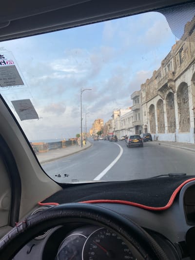 The image depicts a street scene from the perspective of a vehicle's driver, looking forward through a windshield. The location is the city of Beau Fraisier, Algeria.
On the left, a body of water, likely the Mediterranean Sea, stretches to the horizon under a sky with scattered white and grey clouds. Adjacent to the sea is a concrete promenade or sidewalk, bordered by a dark metal railing. Several individuals are present on this promenade; at least three figures are walking together near a lamppost, and others further down appear to be engaged in fishing with rods extending over the water.
The road itself is asphalt, curving gently to the right. A prominent solid white line separates the visible traffic lane from an oncoming or turning lane. Several vehicles are on the road; a black SUV or car with a yellow rear license plate is positioned in the foreground of the traffic flow, with other cars visible further down the road.
On the right side of the road, a continuous line of multi-story buildings with an aged appearance dominates the streetscape. These structures feature a distinct architectural style characterized by large, arched openings at ground level, many of which are partially filled or blocked, and balconies on the upper floors. The facades exhibit signs of wear, including faded paint, crumbling plaster, and exposed stone or brickwork in places. Tall, black streetlights are regularly spaced along the roadside. Another black vehicle is parked alongside the curb near the buildings.
Inside the vehicle, the top portion of a black steering wheel with a worn, stitched leather cover is visible at the bottom of the frame. Above it, a black dashboard is covered with a black mat, accented by a thin red cord outlining its edge. Part of the instrument cluster is visible, showing a speedometer with the needle pointing towards a low speed, likely indicating the vehicle is stationary or moving very slowly. The windshield displays several rectangular stickers on the upper left, some with inverted text, and faint markings or smudges across its surface. In the lower right of the windshield, "RENAULT" text is faintly visible.