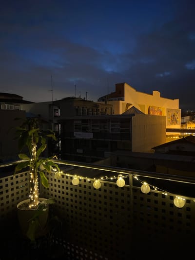 The image depicts a nighttime view from a balcony in Mollet del Vallès, Spain. In the foreground, a potted plant adorned with string lights is situated on the balcony. The balcony railing is also decorated with a string of spherical paper lanterns and small fairy lights. Beyond the balcony, a cityscape is visible with buildings of various heights and architectural styles. Some buildings are illuminated, while others are in shadow. A mural is visible on the side of a larger building. The sky is dark with some visible clouds, suggesting it is evening or night. There are no people visible in the image.