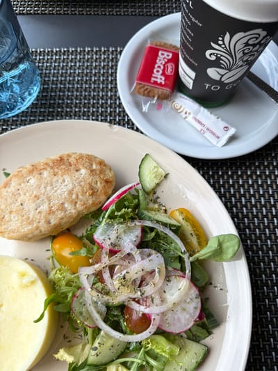 This is a top-down, close-up shot of a breakfast or light meal set on a textured placemat, likely in a hotel or cafe setting. The foreground features a portion of a blue, textured glass, suggesting a drink. Dominating the right side of the frame is a plate of food. On this plate, a light-colored, oval-shaped patty, possibly chicken or fish, is positioned at the bottom. To its right, a vibrant salad is presented, composed of mixed greens, thinly sliced radishes, cucumber rounds, cherry tomatoes (one yellow, one red), and thinly sliced red onion rings, all sprinkled with herbs. A slice of what appears to be cheese or a similar dairy product is partially visible at the bottom right edge of the plate.
To the left of the main plate, a white bowl holds a dark coffee cup or thermos with a decorative white pattern and some text. Adjacent to it are a sugar packet and a single, foil-wrapped Biscoff cookie with its distinctive red packaging. The overall impression is one of a simple, yet appealing meal. The lighting suggests it's daytime. The text visible includes brand names like "Lotus Biscoff" and other labels on the coffee cup and sugar packet. The scene is uncluttered, with the focus on the food and accompanying items. The location context of "la Vila Joiosa, Spain" does not provide any direct visual cues within the image itself, but implies a Mediterranean setting.