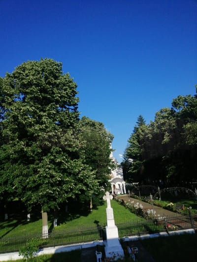 The image depicts a bright, sunny day with a clear blue sky over a church and its surrounding grounds in Horia, Romania. In the foreground, a white cross stands in a grassy area, marked by a low black fence. Beyond the fence, a pathway lined with flowerbeds and benches leads towards the church, which features white walls and a domed roof structure. Large, leafy green trees are prevalent throughout the scene, casting shadows on the well-maintained lawn. The overall atmosphere is tranquil and serene, suggesting a peaceful setting within the town.