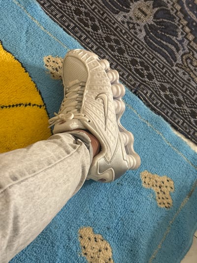 A person wearing light-wash denim jeans is shown from the mid-thigh down, with their right foot adorned with a white Nike Shox TL sneaker. The shoe sits on a multicolored rug with blue, yellow, and tan cloud-like patterns, while a patterned dark blue rug is visible in the background to the left. The location is unknown, and there is no information to suggest that this image was taken in Macedo de Cavaleiros, Portugal.