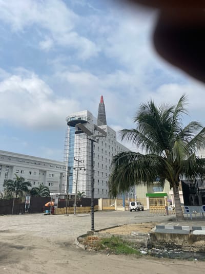 The image depicts an urban landscape in Ikota, Nigeria, under a sky with scattered white clouds. In the foreground, an unpaved dirt and gravel area transitions to a low concrete curb bordering a ditch containing accumulated refuse. A prominent dark utility pole, partially enveloped by scaffolding extending upwards, stands centrally, supporting an arm with a solar panel. To the left of the pole, a small stall with an orange umbrella is visible, beneath which an individual is seated. A light-colored wooden picket fence runs behind the stall.
The midground features a large palm tree partially obscuring the view on the right. The dominant structure is a multi-story white building of modern architectural design, characterized by blue-tinted glass sections and visible exterior scaffolding. This building includes a distinct circular, glass-enclosed upper level and is crowned with a tall, narrow, pointed architectural element topped with a red cap. To the left and slightly behind this, another large, white, multi-story building is visible. A golden metal gate provides access to the main building's compound, where a white SUV is visible behind the gate alongside several flags on poles. To the right of the gate, light-colored outdoor benches are positioned near a building with a green roof.