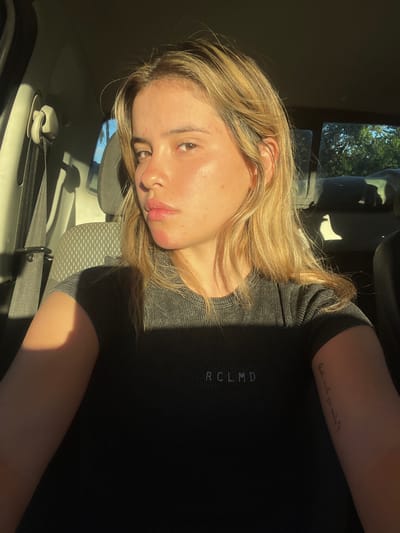 Within a vehicle located in Santa Rosa, Argentina, a young female is captured in a self-portrait. She possesses long, wavy blonde hair and a neutral expression, with her gaze directed towards the camera. Her skin shows minor redness on the chin and cheeks. She is attired in a dark, ribbed, short-sleeved top displaying the white text 'RCLMD' on the chest. A faint, illegible text tattoo is visible on her left forearm. The internal environment of the vehicle includes a gray fabric seat and a seatbelt mechanism on the left side of the frame. Strong, warm sunlight illuminates the left side of her face and torso, creating pronounced shadows on her right side. Through the vehicle's rear window, green foliage is visible against a bright sky, indicating an outdoor setting.