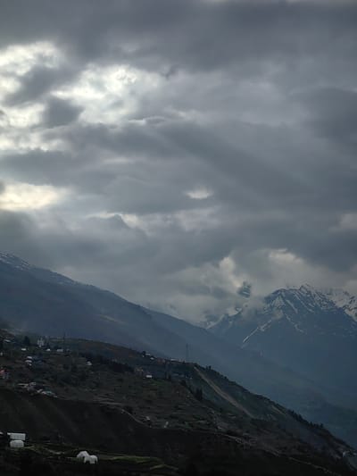 This image captures a dramatic, overcast sky with shafts of sunlight breaking through the thick clouds. Below, a mountainous landscape unfolds. The slopes are covered in green vegetation, dotted with numerous small, white structures that appear to be greenhouses or shelters. Further up the hillside, several buildings are visible, suggesting a small village or settlement. The peaks of distant mountains are snow-capped, contrasting with the darker, lower slopes. The scene is set in Gondhla, India, a location known for its stark beauty and challenging terrain. The overall impression is one of a serene yet powerful natural environment, with hints of human presence adapting to the landscape.