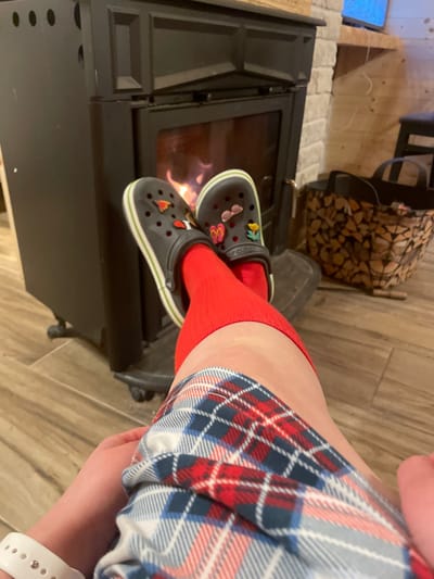 This image, reportedly located in Palež, Montenegro, displays a first-person perspective view of a person's lower body within an indoor setting. The individual's legs are visible from above the knee, clad in a red, blue, and white plaid garment, likely pajamas or lounge pants. Bright red mid-calf socks are worn on both feet, which are inserted into dark gray Crocs featuring light green and white striped soles. These Crocs are accessorized with multiple colorful Jibbitz charms, including a red heart, pink butterfly, and yellow flower, among others. A white wristband, possibly a smartwatch, is visible on the left wrist. The feet are positioned elevated, directly in front of a black, metal wood-burning stove that exhibits active flames behind its glass door. The stove rests on a dark metal floor plate. To the right of the stove, a woven basket filled with split firewood is present. The floor consists of light brown, wood-patterned tiles. The background features a wall segment constructed from light-colored stacked stone, adjacent to a section of light wood paneling. A narrow wooden shelf is visible above the stone wall, and a portion of a lighted screen, possibly a television, is discernible in the upper right corner.