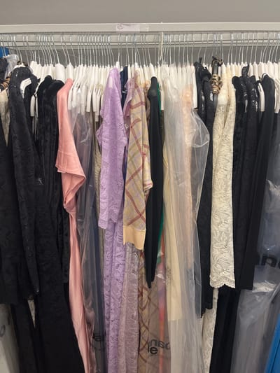 A rack densely packed with clothes on hangers, largely covered in clear plastic garment bags. Predominantly black and lace garments appear, interspersed with various colors and textures like pink, lilac lace, and plaid. The left edge shows the metal rack's structure with a label. The clothing is stacked closely, obscuring individual details.