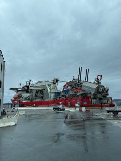 The image depicts a large, specialized vessel, identified as "Subsea 7 SEVEN NAVICA DOUGLAS," docked alongside a concrete quay in Bergen, Norway. The vessel is predominantly red and grey, with various industrial structures and equipment mounted on its deck, including cranes, pipes, and complex machinery. A black car is parked nearby on the wet asphalt surface, indicating recent or ongoing rain. A white trailer is positioned beside the vessel at the dock, suggesting a logistical operation. The sky is overcast, typical of Bergen's coastal climate. A corner of a grey, multi-story building is visible in the lower left corner of the frame. The wet pavement reflects the overcast sky and surrounding structures.