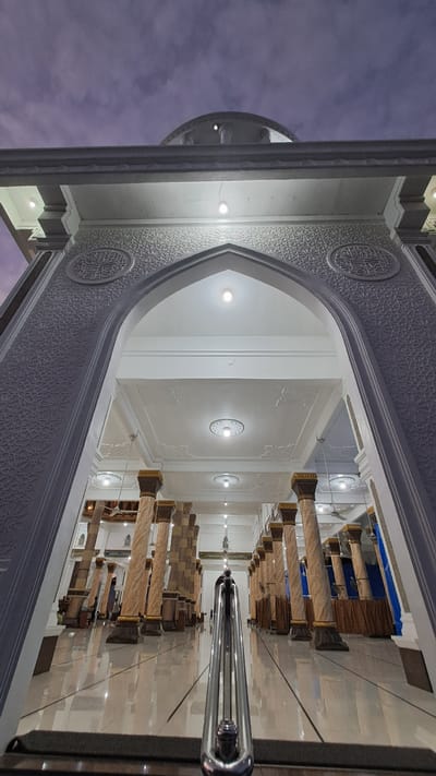 The image shows the entrance to a mosque in Lhokawe Teungoh, Indonesia. The viewpoint is low, looking up through a large arched entryway. The mosque has a white facade with intricate geometric carvings and Arabic calligraphy. Inside, the mosque is supported by numerous tall columns with marble-like shafts and dark brown bases and capitals. The ceiling is white with recessed lighting fixtures. The floor is highly polished and reflective, showing the reflections of the columns and lights. The sky visible above the mosque entrance is a twilight sky with some clouds. There are a few people visible inside the mosque, indistinctly. A metal railing is prominent in the foreground, leading up towards the entrance.