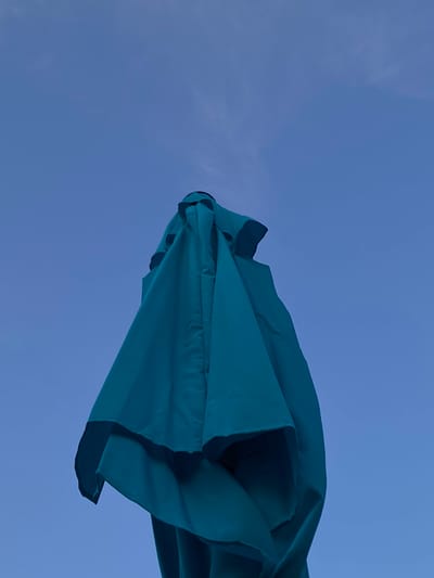 A close-up, low-angle shot shows a closed teal-colored patio umbrella against a deep blue sky with wispy white clouds. The umbrella is the primary subject, occupying the majority of the frame. Its fabric is gathered and bunched, creating folds and shadows that give it a sculptural appearance. The umbrella's top, where the fabric meets the pole, is visible, revealing a dark rim. The sky is a vast expanse, with the blue deepening towards the top of the image. The lighting suggests it is either early morning or late afternoon, as the sky is not bright white and the umbrella is not brightly illuminated. The image was taken in the city of Arandjelovac Municipality, Serbia.