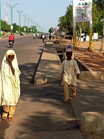 The image captures a street scene in Wuro Dole, Nigeria, under a clear, bright sky. In the foreground, two children are walking on a paved sidewalk. The child on the left, facing away from the camera, wears a pale yellow headscarf and robe, with brown sandals. The child on the right, facing towards the camera, is a boy in a pale cream-colored tunic and pants, also wearing sandals. Behind the children, a street stretches into the distance, lined with trees and lampposts with solar panels. Vehicles, including motorcycles and cars, are visible on the road, with people walking and riding. A prominent signpost with text, partially obscured, stands to the right of the children. The environment suggests a typical urban or semi-urban setting in Nigeria.