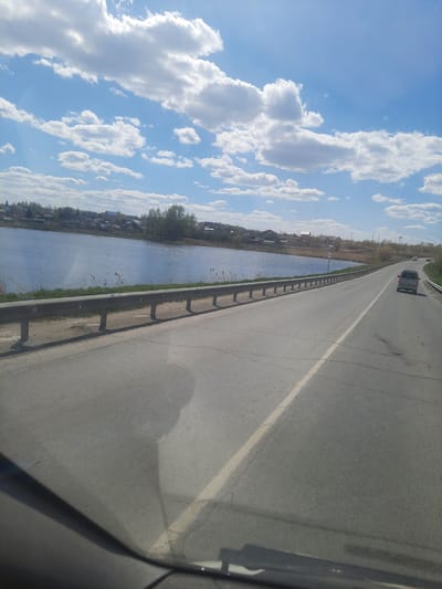 The image depicts a view from a vehicle traveling on a road in Научный Городок, Russia. The road runs parallel to a body of water with vegetation and buildings visible on the opposite shore. A guardrail separates the road from the water. The sky is blue with scattered clouds. A vehicle is ahead on the road.