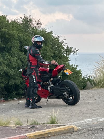A motorcyclist wearing a red and black Dainese racing suit and helmet stands next to a red sport motorcycle. The motorcyclist is facing away from the camera and towards a body of water, possibly the sea. The motorcycle has a yellow license plate. The scene is set outdoors, with green foliage and the sea in the background. The ground is paved. The image is likely taken in a coastal area in the Mate Asher Regional Council, Israel, given the license plate and the vegetation.