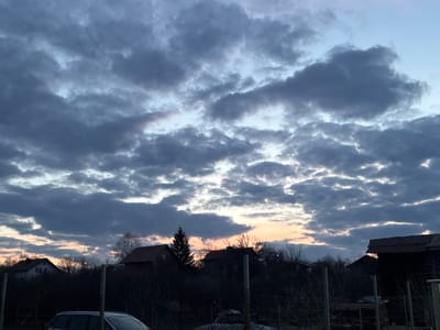 The image depicts a rural or semi-urban landscape in Zidartsi, Bulgaria, under twilight conditions, likely dawn or dusk. The sky is largely overcast with extensive, dark grey cloud cover, fragmented to reveal patches of lighter blue sky and a band of orange-pink light near the horizon. The mid-ground features a dark silhouette of residential structures, specifically houses with visible pitched, tiled roofs, interspersed with bare deciduous trees and one prominent coniferous tree. In the immediate foreground, a chain-link fence or similar barrier is partially visible, obscuring the lower portion of a light-colored vehicle, only its upper body and roofline discernible. Multiple dark, vertical fence posts are visible across the foreground. No individuals or discernible activities are present.