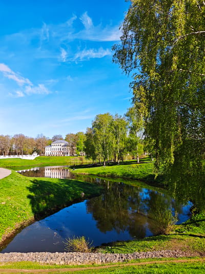 The image presents a landscape scene in Uglich, Russia, featuring a body of water reflecting a classical-style building with white columns. The building, likely a historical landmark, sits amidst a park-like setting with trees. The water's surface mirrors the building's façade and surrounding trees. A stone wall or embankment defines one edge of the water feature, while grassy banks and foliage surround the water. The sky is blue with wispy clouds, suggesting a clear day. A large tree with green leaves is prominent on the right side of the image. The scene conveys a sense of serenity and historical importance.