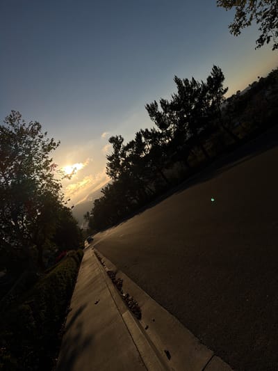 The image shows a street scene in Aliso Viejo, California, at sunset. The sun is low in the sky, casting a warm glow and long shadows. The road is asphalt, and there is a concrete sidewalk with a drainage channel along the side. Dense trees with dark foliage line the street, and a few buildings are visible in the distance on a hillside. The sky is a gradient of blue and yellow, with some scattered clouds. A single car is parked on the side of the road.