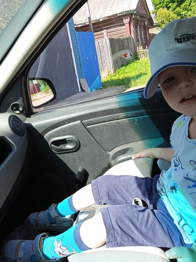 The image shows a young child seated in the passenger side of a car, looking towards the viewer. The child wears a light-colored baseball cap with the Russian text "ЧЕРНО МОРЕ" (Black Sea) visible on the brim, a light blue t-shirt with dark blue patterns, dark blue shorts, bright turquoise socks featuring a cartoon design, and grey sandals. Their face is partially visible, with a neutral or slightly open-mouthed expression.

The car's interior details include a grey plastic door panel, a window, and a side mirror reflecting a dirt road and a distant red-roofed building. Bright sunlight streams into the car, creating distinct shadows on the door and the child's lap.

Outside the car window, the scene reveals a rural or suburban setting in Teykovo, Russia. To the left, there's a blue corrugated metal fence and a dark building. Further along, a traditional Russian wooden house with intricately carved window frames and a gabled roof is prominent. A wooden picket fence runs in front of the house, with green grass and bushes flourishing around it. A white cartoon cat sticker holding a red heart is visible on the car windowpane, partially overlapping the exterior scene.

The bright, clear light indicates it is daytime, likely a sunny and warm day. The scene captures a momentary stop or travel through a residential area, showcasing local architecture.