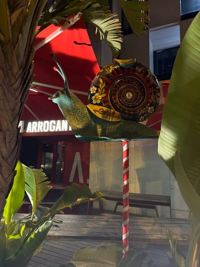 An outdoor, nighttime scene in Eivissa, Spain, features a large, decorative snail sculpture as the focal point. The sculpture, with its iridescent green and gold body and a highly ornate shell adorned with floral motifs and intricate spiral patterns in red, gold, black, and white, is mounted on a red and white striped pole. In the background, a building with illuminated white lettering that reads "1 ARROGANT" is visible behind a red awning. There are large green leaves from tropical plants framing the scene, casting shadows. Wooden benches are visible in the mid-ground, suggesting an outdoor seating area. The lighting is artificial and casts a warm glow on the scene.