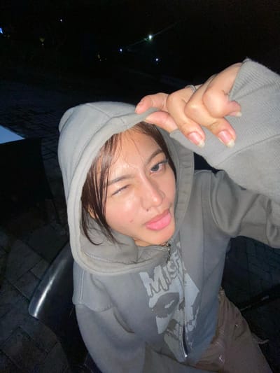 A young Asian woman is captured in a close-up, self-taken photograph, likely at night given the dark, indistinct background. She is wearing a grey hooded sweatshirt, which she has pulled up to partially cover her head. Her left eye is closed in a wink, and her tongue is sticking out from the side of her mouth. Her right hand is raised, with fingers extended and touching the edge of the hood. The woman has a piercing on her lower lip and appears to be wearing a ring on her left ring finger. The lighting suggests an artificial light source, possibly from a phone or camera, illuminating her face and upper body. The background is dark and blurry, with a few faint lights visible, hinting at an outdoor or dimly lit indoor environment. Without further information or context, it is not possible to determine the specific location within Mekar Bakti, Indonesia.