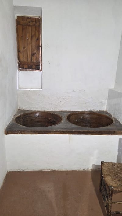 The interior of a room in Altura, Spain, is shown. There is a rectangular opening in the wall at ground level covered with wooden planks. A vertical, decorative feature consisting of two large, circular indentations is positioned against the wall. A portion of a chair with a straw seat is visible in the upper right corner. The walls and floor are white, with one wall painted brown in the upper right.