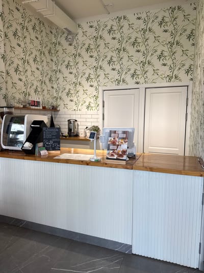 A high-angle, slightly tilted shot shows the interior of a cafe or shop with a modern and nature-inspired design. The walls and ceiling are covered in a white wallpaper with a repeating pattern of green bamboo stalks and leaves. A wooden counter runs across the foreground, with a white, ribbed wall paneling behind it. On the counter, a digital menu display shows images of pastries. To the left of the menu, a small chalkboard sign is visible with handwritten text in Russian. Among the visible text are the words "Лимонады" (Lemonades) and "Моти" (Mochi), along with prices like "179 руб" (179 rubles). Behind the counter, there are shelves with glass jars containing various goods, possibly coffee beans or spices. A coffee machine and other kitchen appliances are also visible. The floor in the lower right corner of the image is made of dark gray tiles with white veining. The scene appears to be indoors, with artificial lighting. No people are visible in the frame. The overall impression is of a clean, stylish, and possibly upscale establishment.