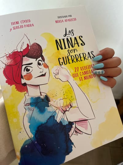 A woman's hand with blue painted nails, one adorned with floral designs, holds a book titled "Las Ninas son Guerreras" (Girls are Warriors). Subtitled "27 Rebels who Changed the World", the book features an illustration of a woman with a red headscarf, winking and holding up a clenched fist. The cover also credits Irene Civico and Sergio Parra as authors, and Nuria Aparicio as the illustrator.