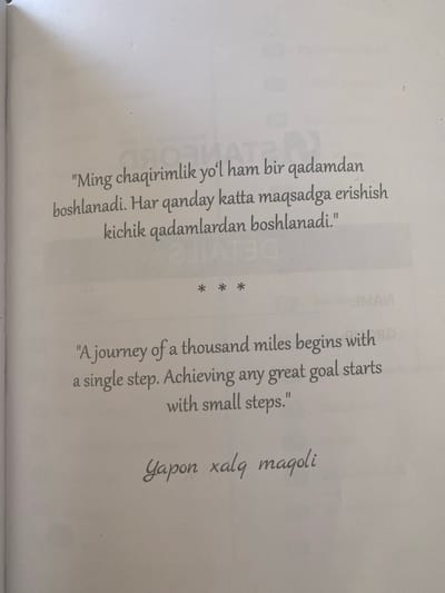 The image shows a page from a book with text in both Uzbek and English. The Uzbek text, which appears to be a quote, is centered on the upper half of the page and reads: "Ming chaqirimlik yo'l ham bir qadamdan boshlanadi. Har qanday katta maqsadga erishish kichik qadamlardan boshlanadi." Below this, there are three asterisks centered. The English text, also a quote, is centered on the lower half of the page and reads: "A journey of a thousand miles begins with a single step. Achieving any great goal starts with small steps." At the very bottom of the page, in a cursive font, is the Uzbek phrase "Yapon xalq maqoli." There are no people, specific actions, or interactions depicted. The environment is that of a book page, with faint, out-of-focus text in the background that appears to be from a different page or overlay. There is no information or visual evidence within the image that connects it to the city of Arbagish, Uzbekistan.