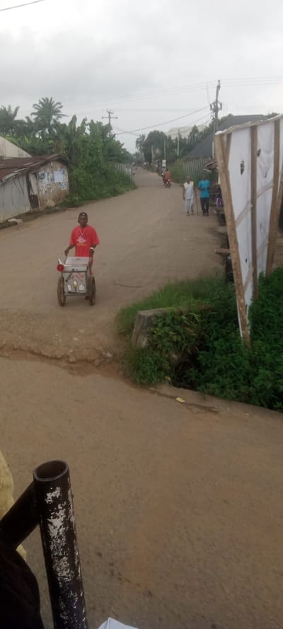 This image depicts an overcast daytime scene on an unpaved road in Ezinifite, Nigeria. In the left midground, a male wearing a red t-shirt and a dark cap pushes a white, two-wheeled vendor cart, which features text including "SPECIAL ICE CREAM" and "HYDRATION IS KEY." In the right midground, two males walk away from the viewer; one, dressed in a white tank top and light trousers, uses a walking stick, while the other wears a blue t-shirt and dark trousers. Further down the road, a motorcyclist is visible, also traveling away from the camera. The road itself is wide, unpaved or graveled, and appears to have visible ruts and worn patches. Both sides of the road are bordered by dense green vegetation, including numerous palm trees and bushes. Utility poles with overhead power lines are visible along the road. On the left, simple structures with corrugated metal roofs are present, some displaying faded posters. On the right, a makeshift wooden structure with white paneling stands near a concrete culvert or drainage structure overgrown with vegetation. The sky is uniformly gray and overcast. A dark, weathered metal pipe object is partially visible in the lower left foreground, suggesting the image was taken from a vehicle or similar elevated perspective.